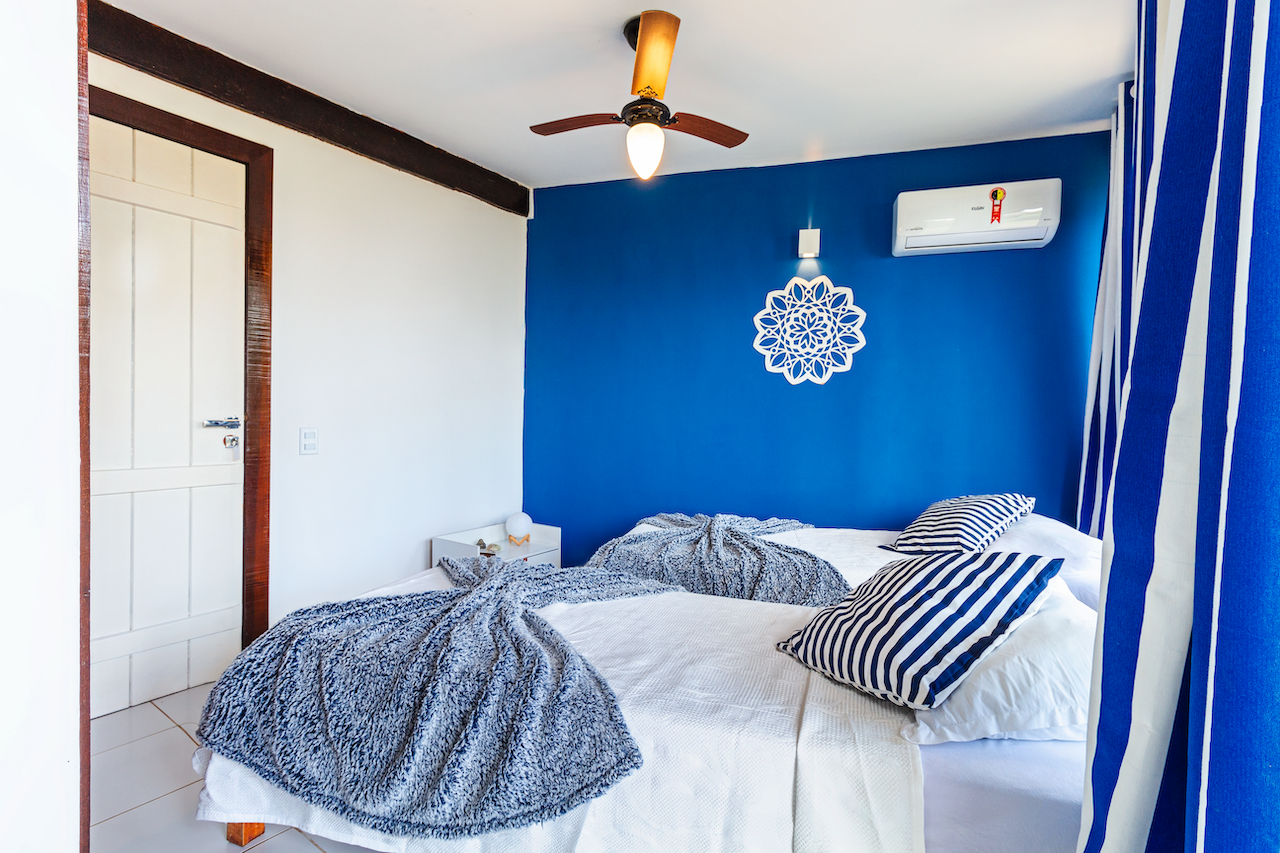 A bedroom with two unmade beds, a blue accent wall with a white decorative wall piece, striped pillows, and curtains. There is a ceiling fan with wooden blades and an air conditioning unit on the wall.