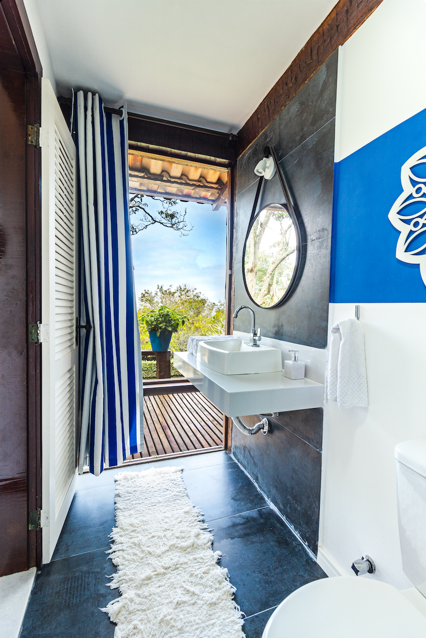 Bathroom with a white sink, black and white round mirror, white towel, and a view to an outdoor balcony with a plant in a blue pot and trees outside.