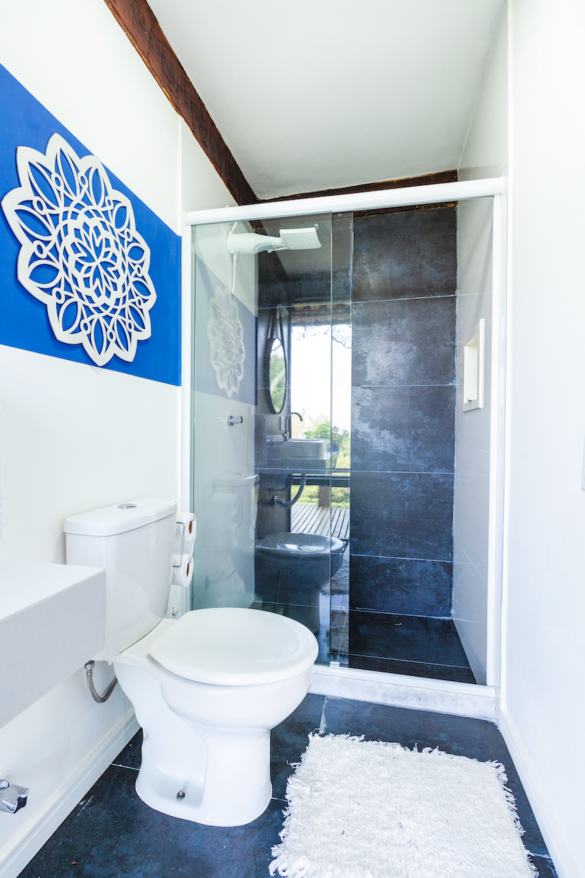 A small, modern bathroom featuring a white toilet, a white sink, a black tile shower with a glass door, a rain showerhead, and a decorative blue wall art piece. There's a white fluffy rug in front of the toilet and the bathroom has dark flooring.