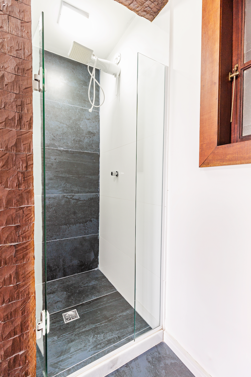 View of a modern shower with gray tiles, a white wall, and a glass door, adjacent to a red brick wall and a wooden window frame.