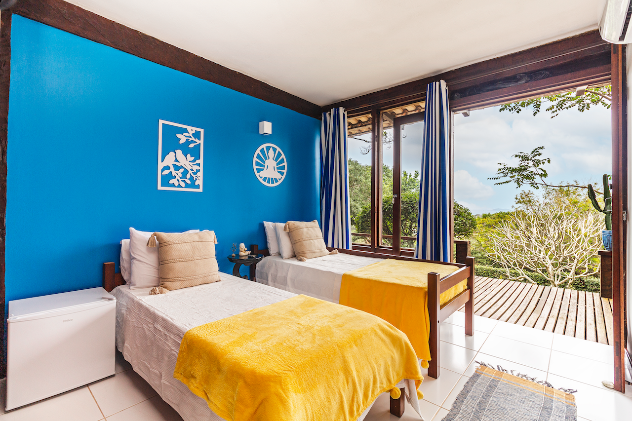 A bright hotel room with two single beds, yellow blankets, and beige pillows, featuring a blue accent wall with decorative wall art, a small black side table, and a large window with blue and white striped curtains that opens to a wooden balcony with