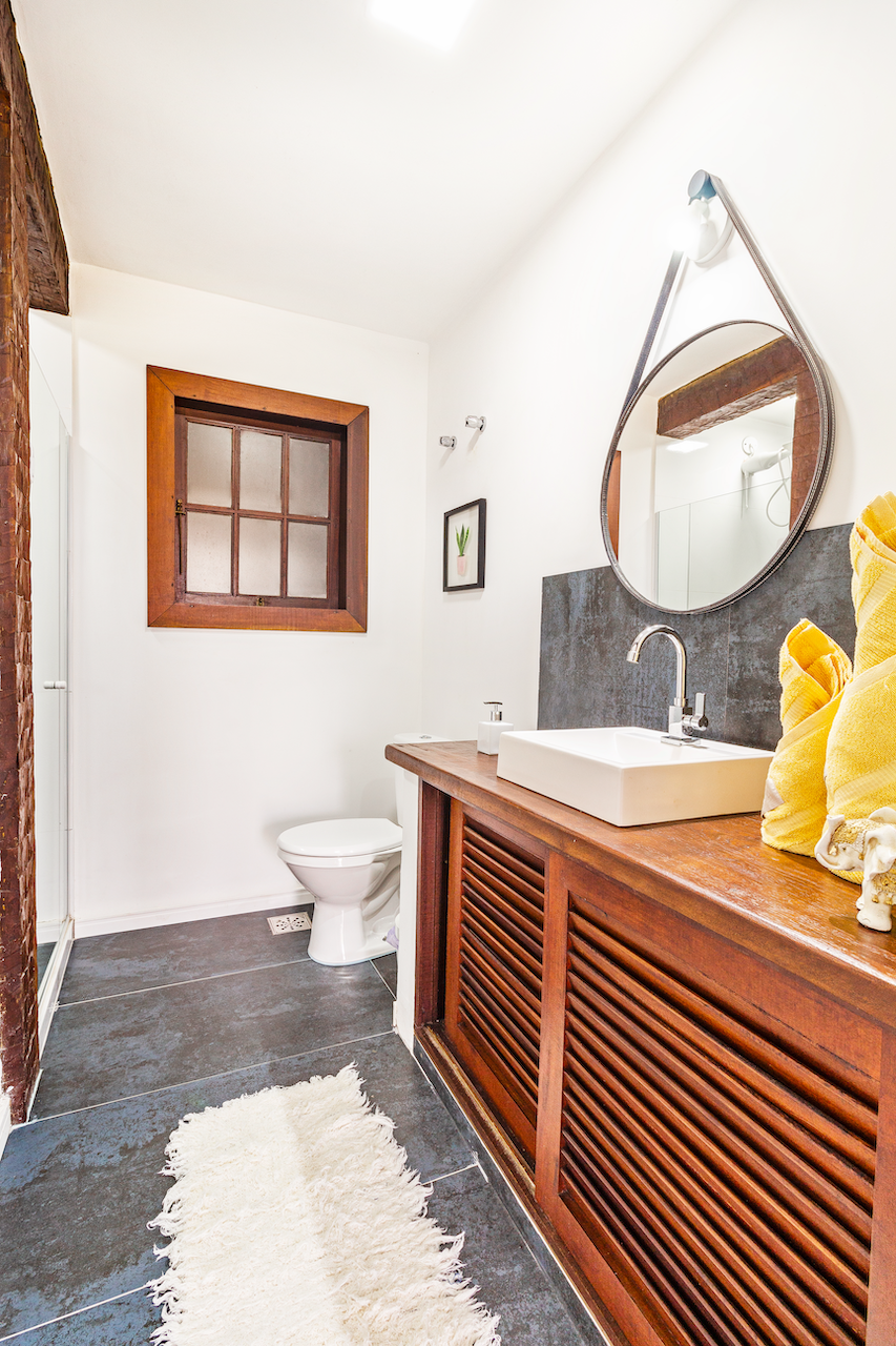 Bathroom with white walls, wooden window frame, and gray tile floor. There is a white toilet and a wooden cabinet with a white sink on top. A round mirror is mounted above the sink, and a small framed plant art is on the wall. Towels and decorative s