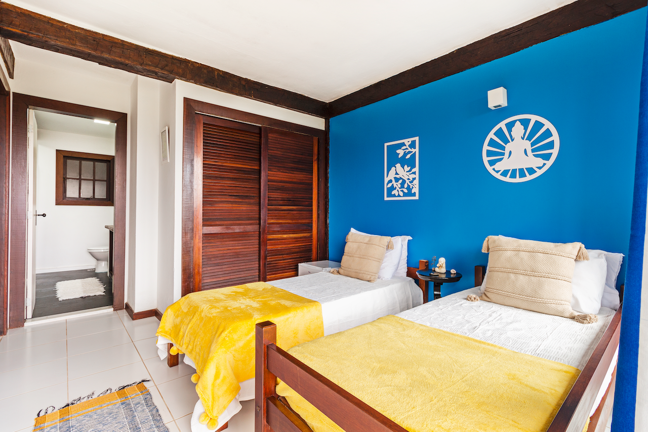 A bright bedroom with two single beds covered in yellow blankets, a blue accent wall with decorative wall art, and a bathroom door visible in the background.
