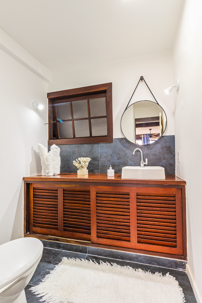 Bathroom with a wooden vanity, a small sink, a round mirror hanging on the wall, and a window with wooden frame. There is a white toilet and a white rug on the dark tile floor, with decorative items on the vanity.
