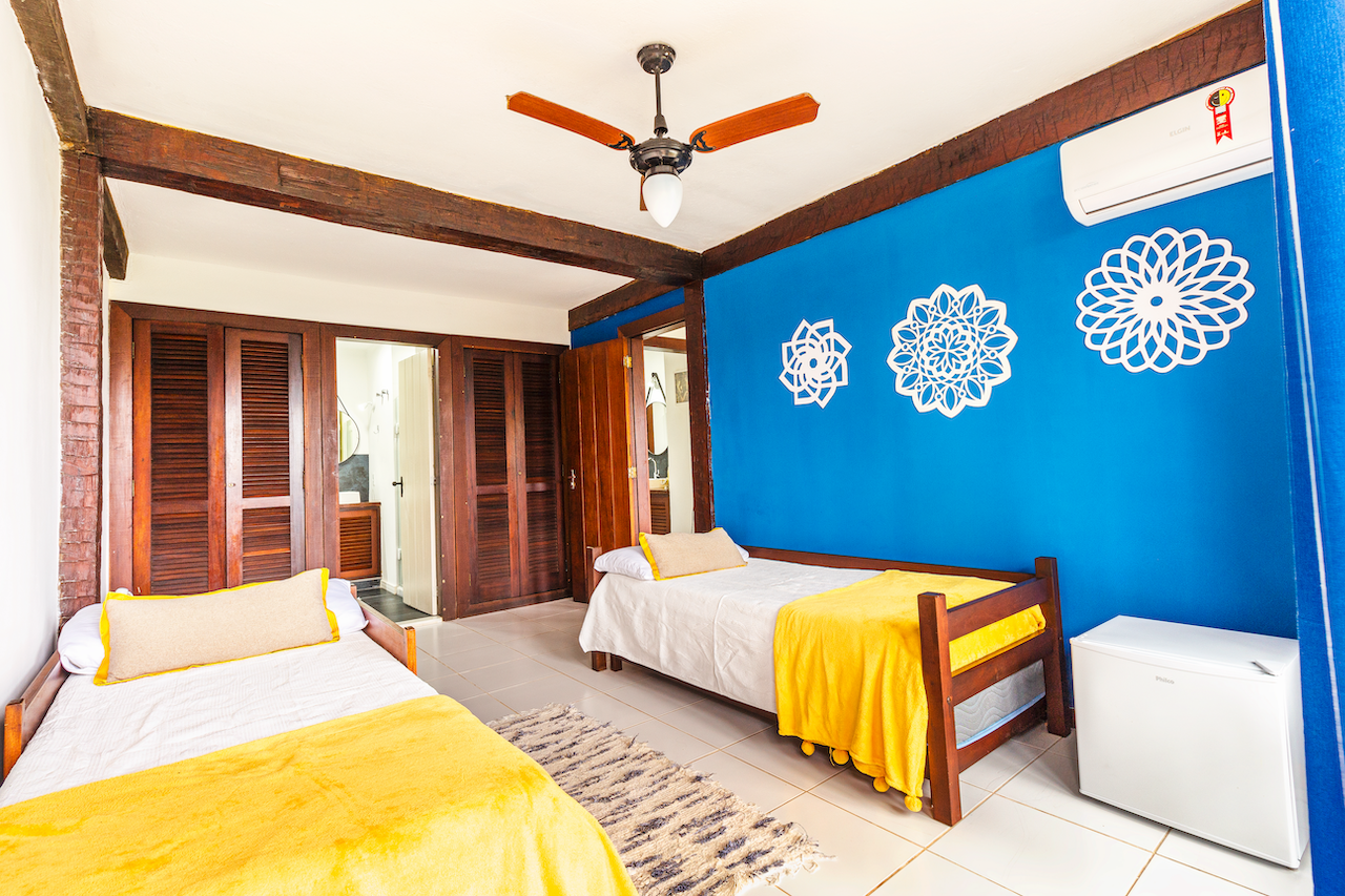 A bedroom with two single beds with yellow blankets and pillows, wooden furniture, a blue accent wall with white decorative wall decals, a ceiling fan, air conditioner, and a small white refrigerator.