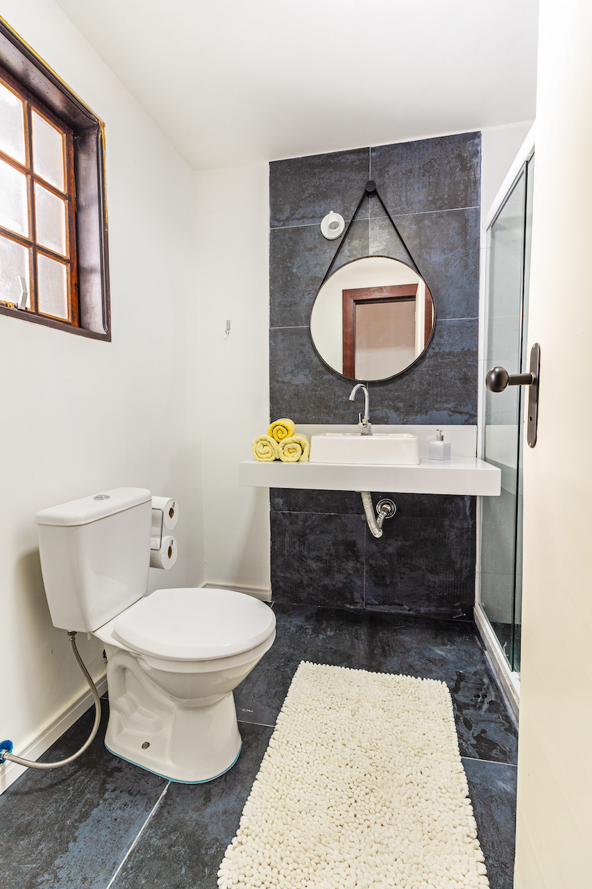 Bathroom with white walls, window, toilet with double toilet paper rolls, black tiled floor, white shaggy rug, floating sink with a mirror above, yellow towels on the sink, and a sliding shower door on the right.