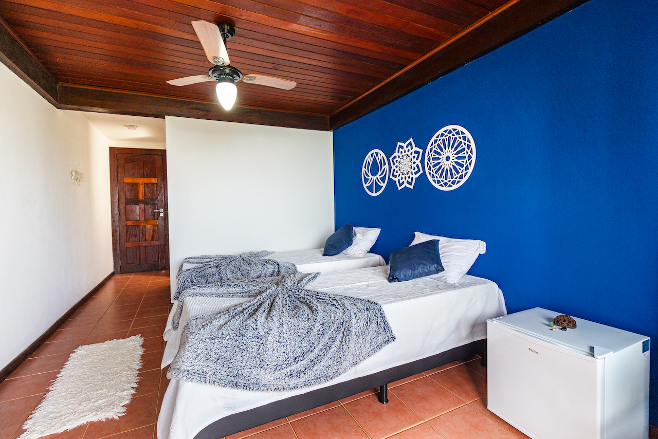 A bedroom with two beds, a blue accent wall with white decorative wall hangings, a wooden ceiling with a ceiling fan, and a white mini fridge.