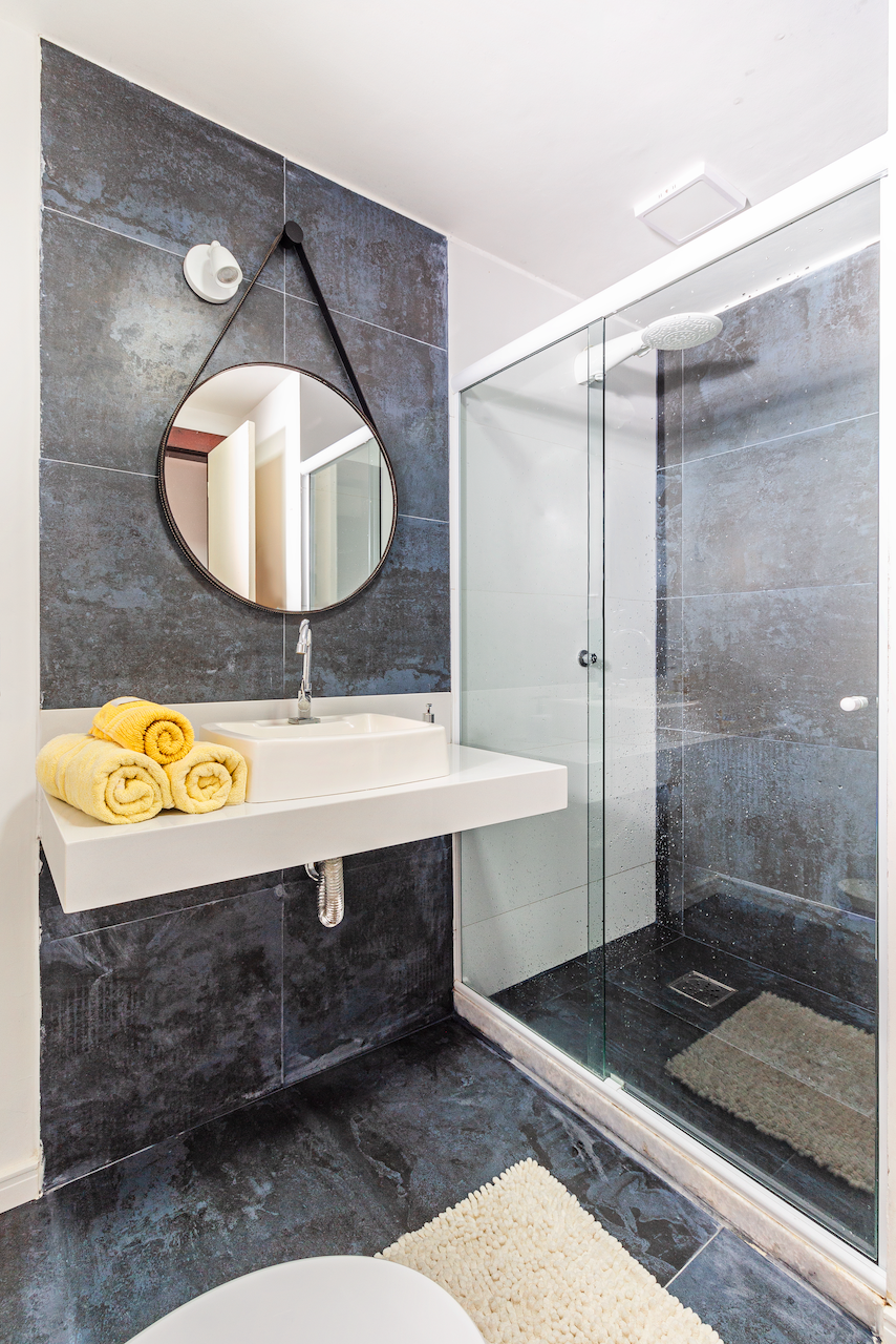 Modern bathroom with dark gray tiled walls and floor, a white sink mounted on a glazed shower wall, a round mirror hanging from a black strap, yellow rolled towels, a glass shower enclosure with a rainfall showerhead, and a beige bath mat.