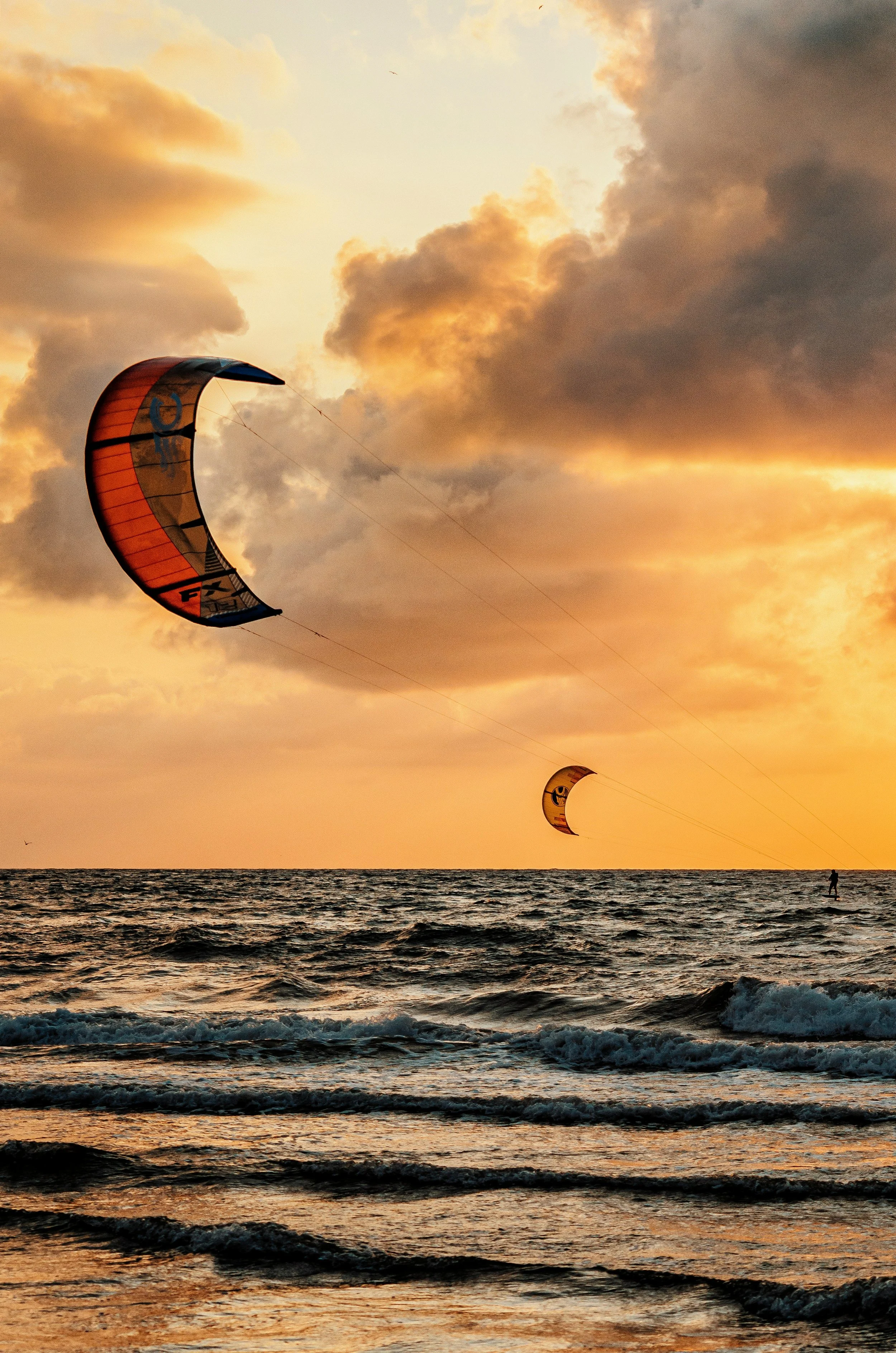 Two people kite surfing at sunset over the ocean, with orange and yellow clouds in the sky.