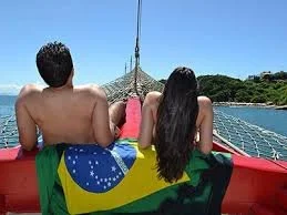 Two people sitting on a boat with a Brazilian flag, facing away and looking at the water, under a clear blue sky.