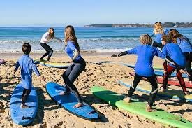 Group of children in blue shirts on surfboards at the beach with an adult instructor, practicing surfing skills near the shoreline.