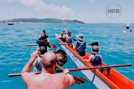 A group of people in a boat with paddles on a large body of water, some wearing hats and sunglasses, with land in the distance.