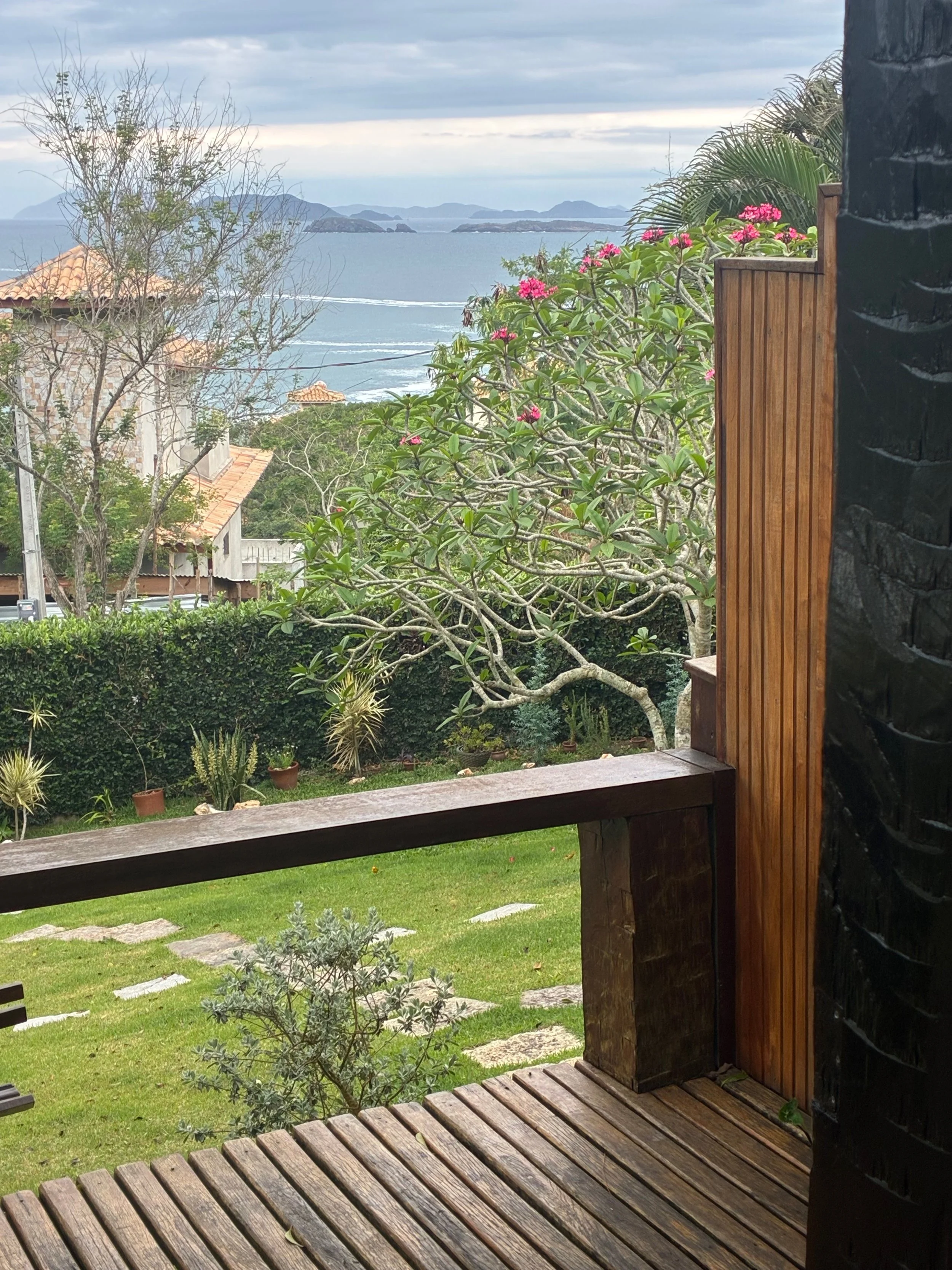 View from a wooden deck overlooking a lush garden with green grass, potted plants, and trees, with a scenic ocean and islands in the distance under a cloudy sky.
