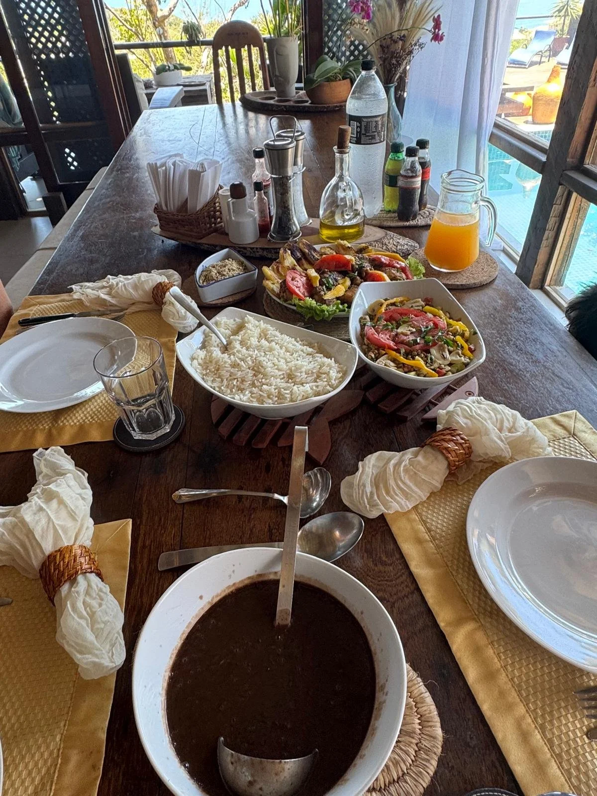 A dining table set with various dishes including rice, salad, and a dark soup, along with drinks such as orange juice and bottled beverages. The table is partially set with plates, utensils, and napkins, and is indoors near a window with a view of a 