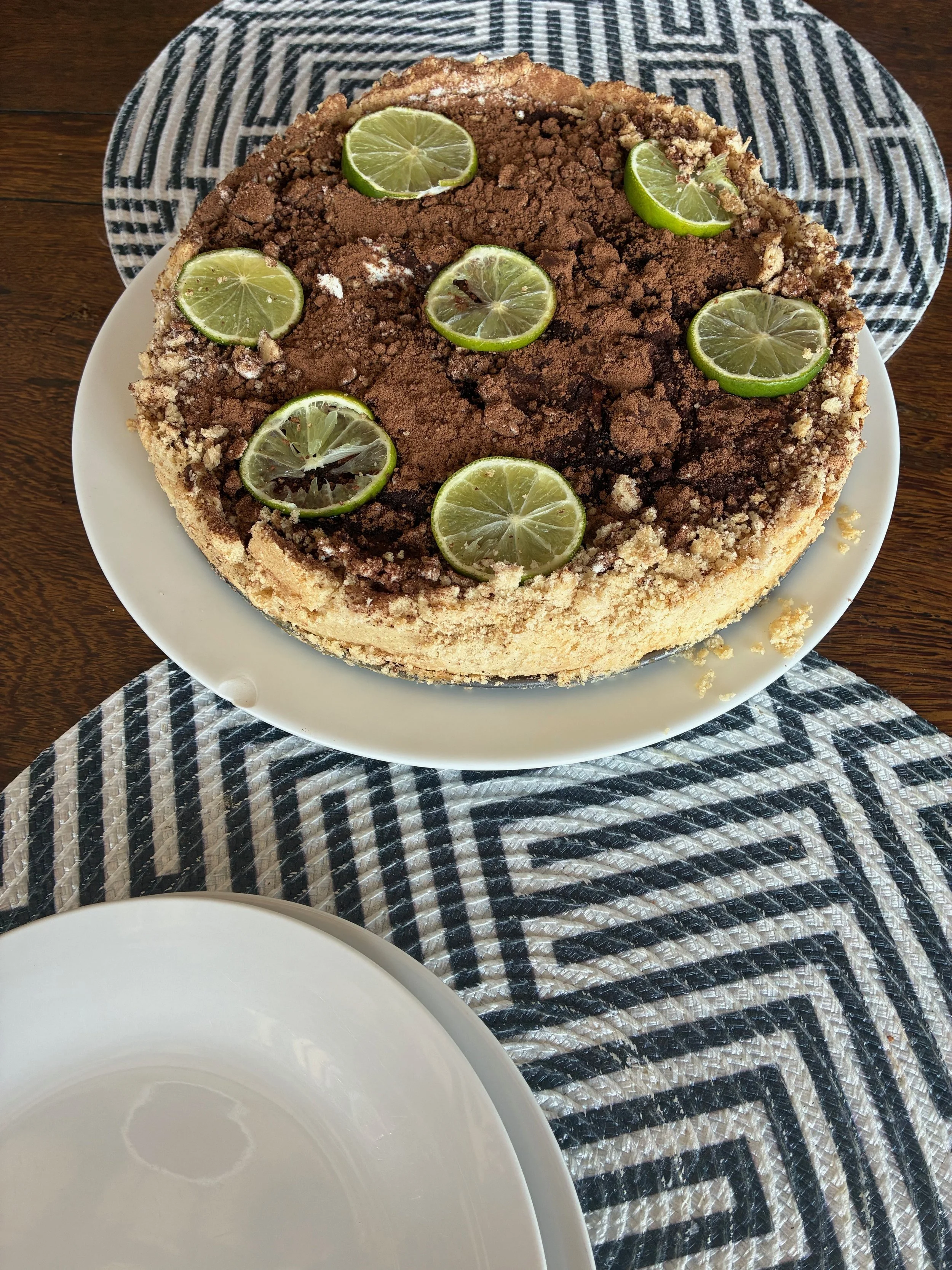 Chocolate cake topped with lime slices, on a white plate, placed on a black and white patterned tablecloth.