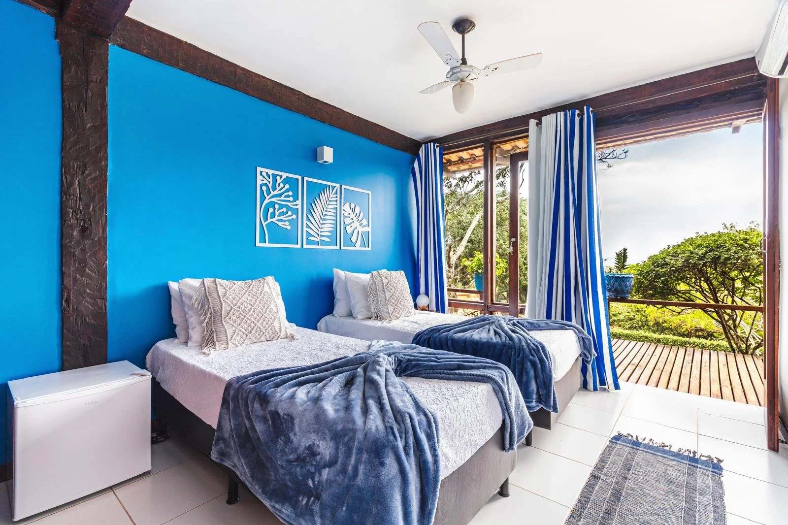 A bedroom with two twin beds pushed together, each with white bedding, beige pillows, and blue blankets. The wall behind the beds is painted blue with white decorative wall art. There is a small white mini refrigerator beside the beds. Large glass do