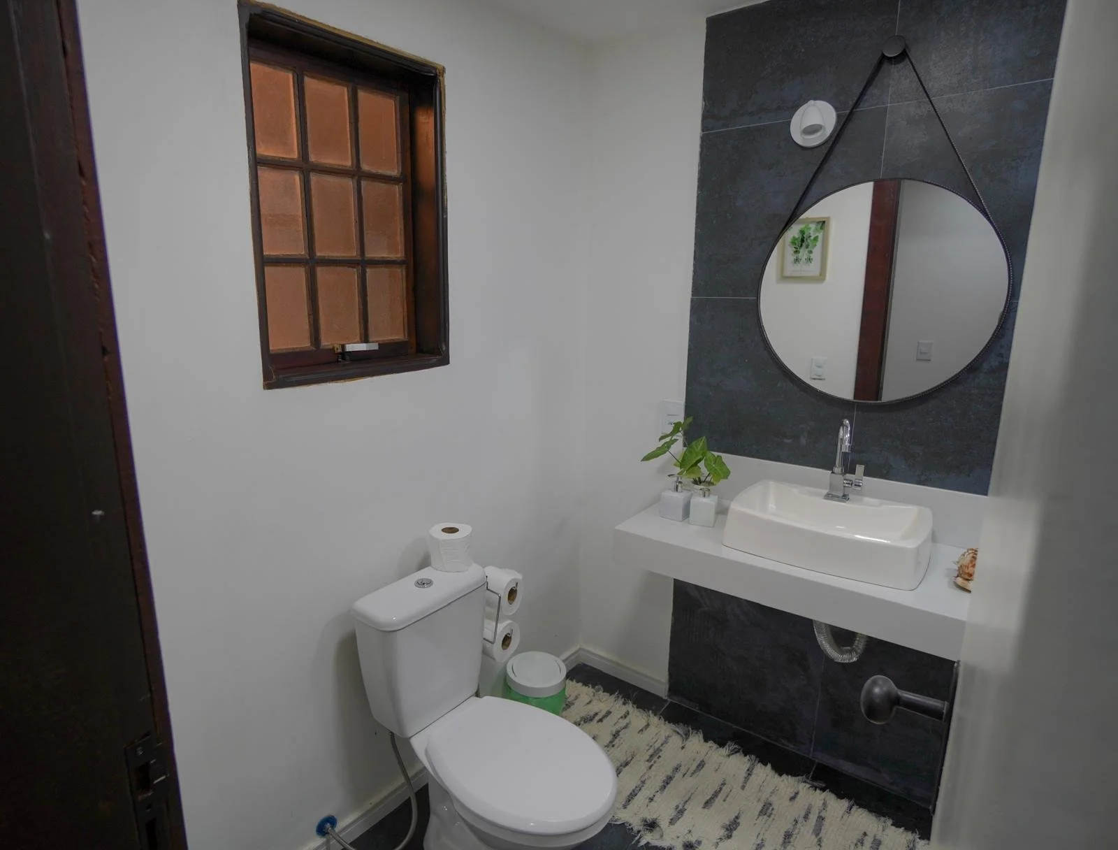 A bathroom with a toilet, a sink with a mirror above it, and a small window. The mirror is round, hanging by a strap, and there's a plant and decorative items on the sink counter. The bathroom has white walls and dark tiles behind the mirror.