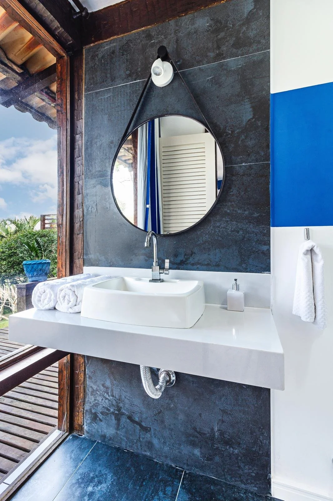 Modern bathroom with white square vessel sink, silver faucet, round mirror with black leather strap, dark textured wall, white towel, soap dispenser, and a large window showing a wooden deck and greenery outside.