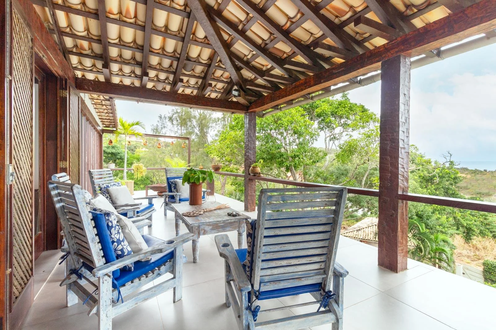 Outdoor balcony with wooden chairs and a table, green trees in the background, and a tiled roof visible in a sunny setting.