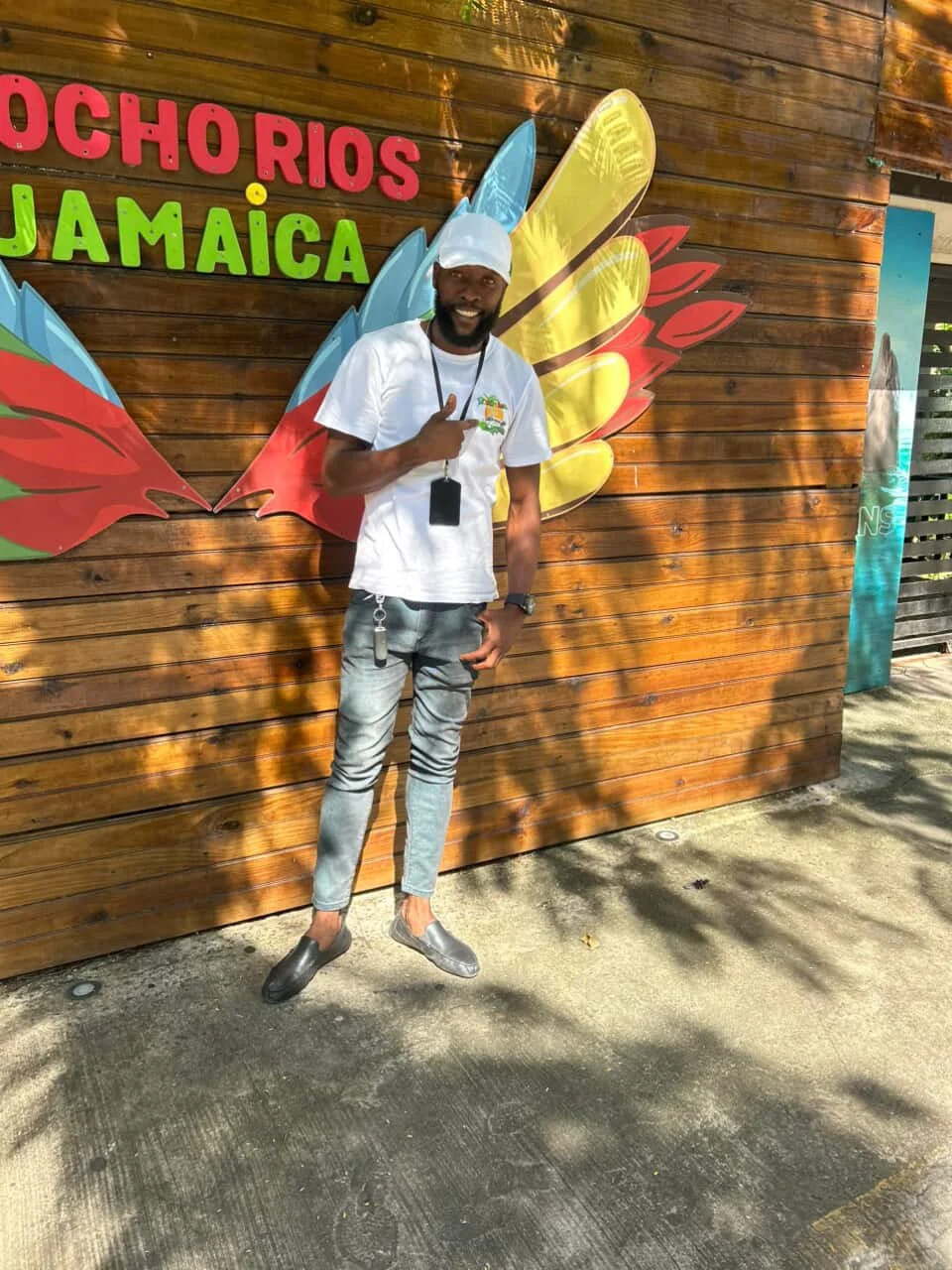 A man standing in front of a wooden wall with colorful signage that reads 'Ocho Rios Jamaica'. He is smiling, wearing a white t-shirt, light jeans, gray slip-on shoes, a white cap, and has a lanyard with cards around his neck. He is pointing his right thumb up and has a keychain hanging from his pocket.