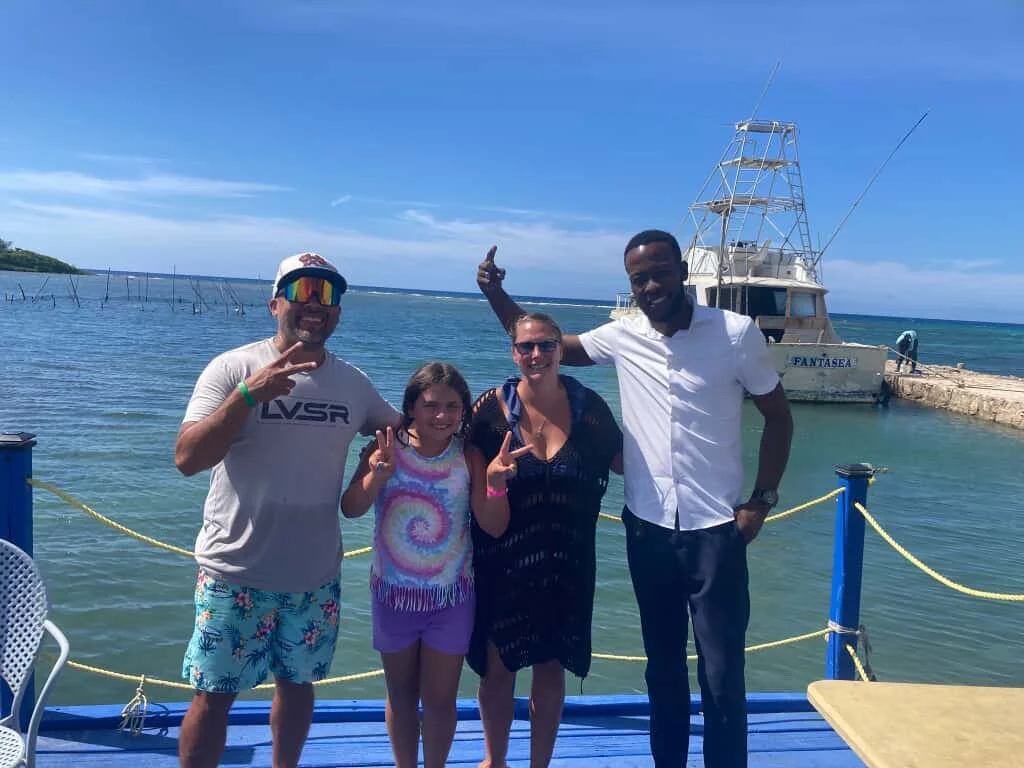 Four people, two men and two women, standing on a dock near a boat with the ocean and a clear blue sky in the background, posing for a photo.
