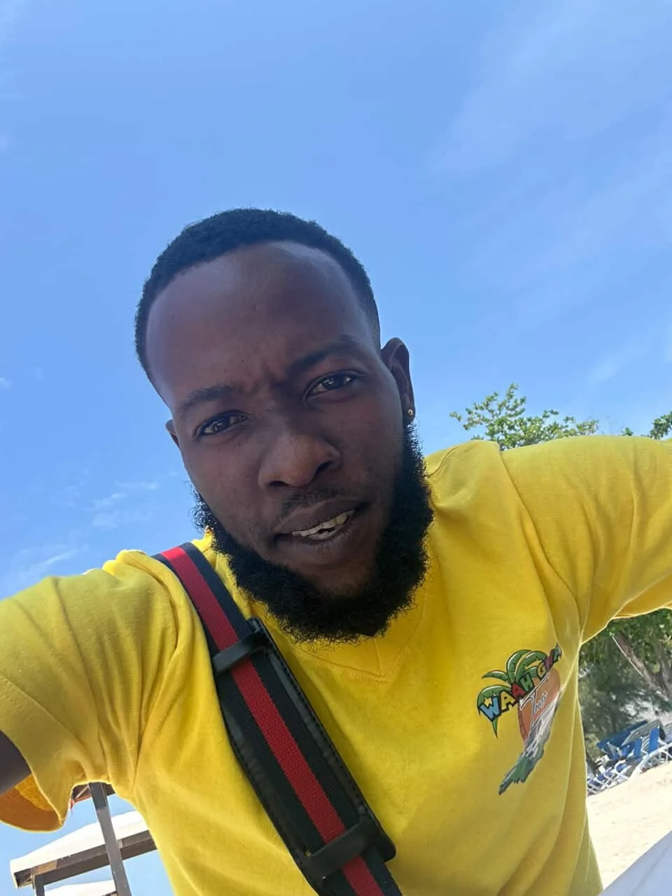A young man with a beard wearing a yellow T-shirt outdoors on a bright day, with blue sky and some trees in the background.