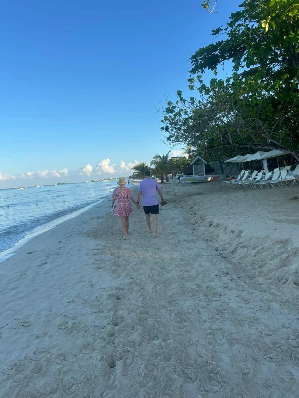 A couple walks hand in hand along a sandy beach, with the woman wearing a sunhat and floral dress and the man wearing a purple shirt and dark shorts. The beach has lounge chairs and umbrellas on the right, and there are trees and a small building in 