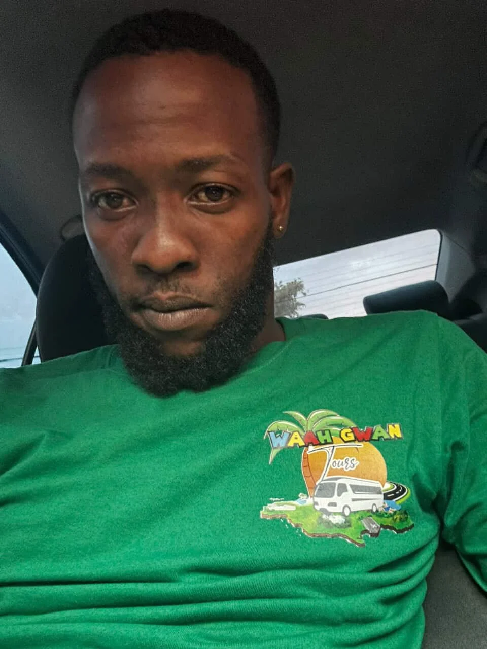 A man sitting in a car, wearing a green T-shirt with a colorful logo that says 'WAAH GWAN Tours,' featuring a palm tree, a tour bus, and a map of the U.S.