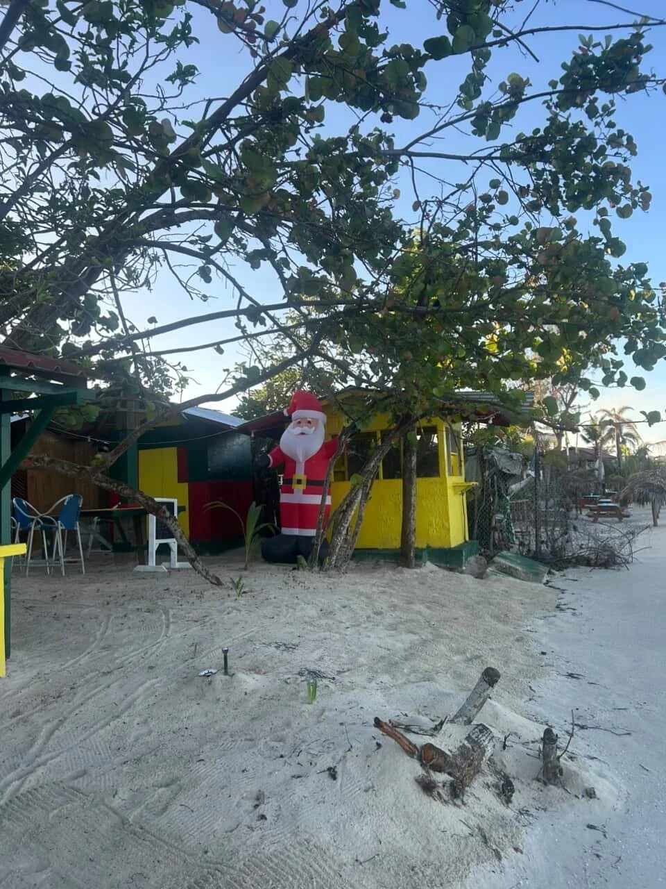 A small colorful beach hut with a Santa Claus inflatable figure beside it, surrounded by sandy ground and trees.