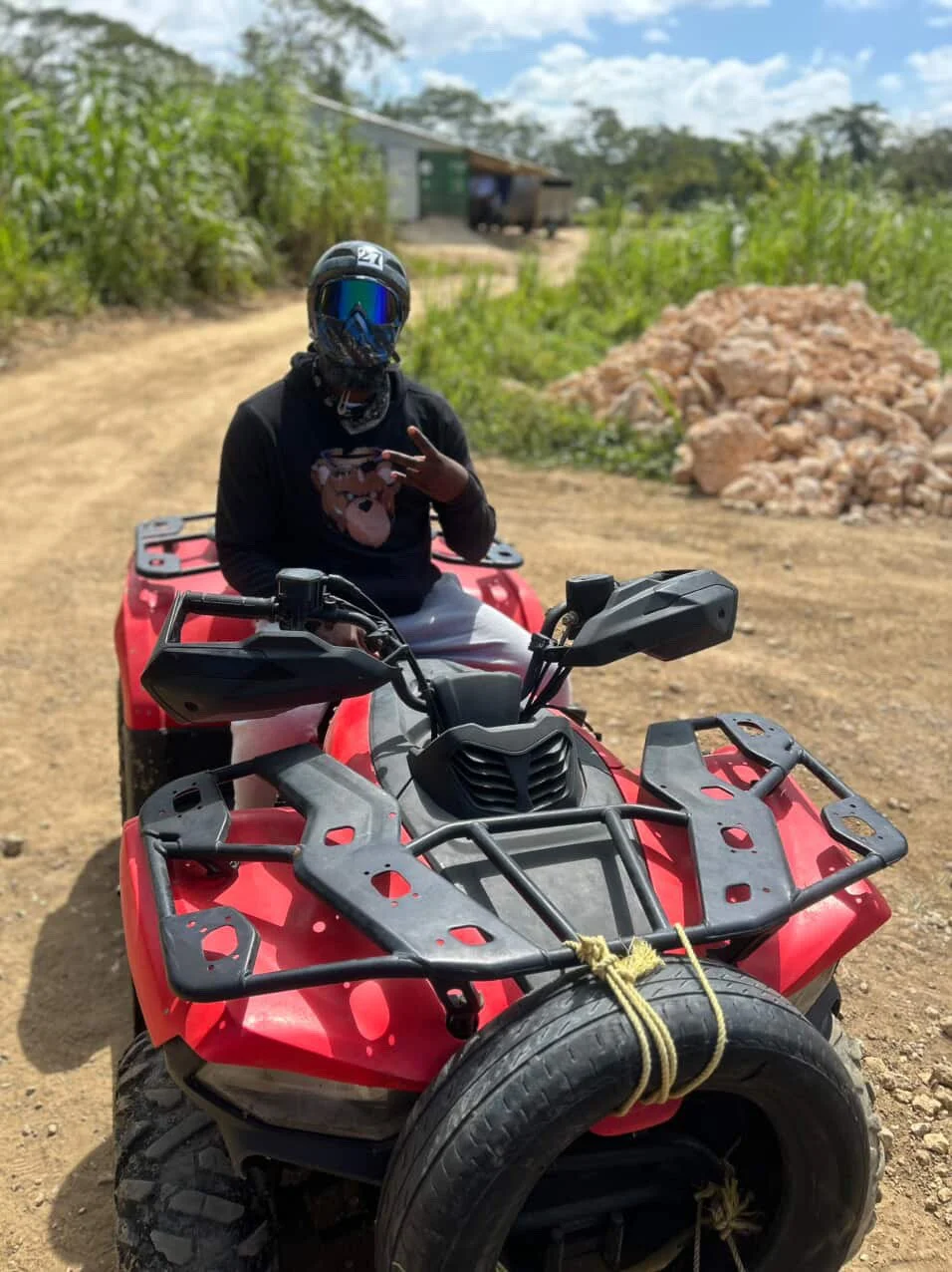 Person sitting on a red all-terrain vehicle (ATV) on a dirt road, wearing a black helmet with blue visor, black hoodie with a bear graphic, and making a peace sign with their right hand. In the background, there are green bushes, a small building, and a pile of rocks.