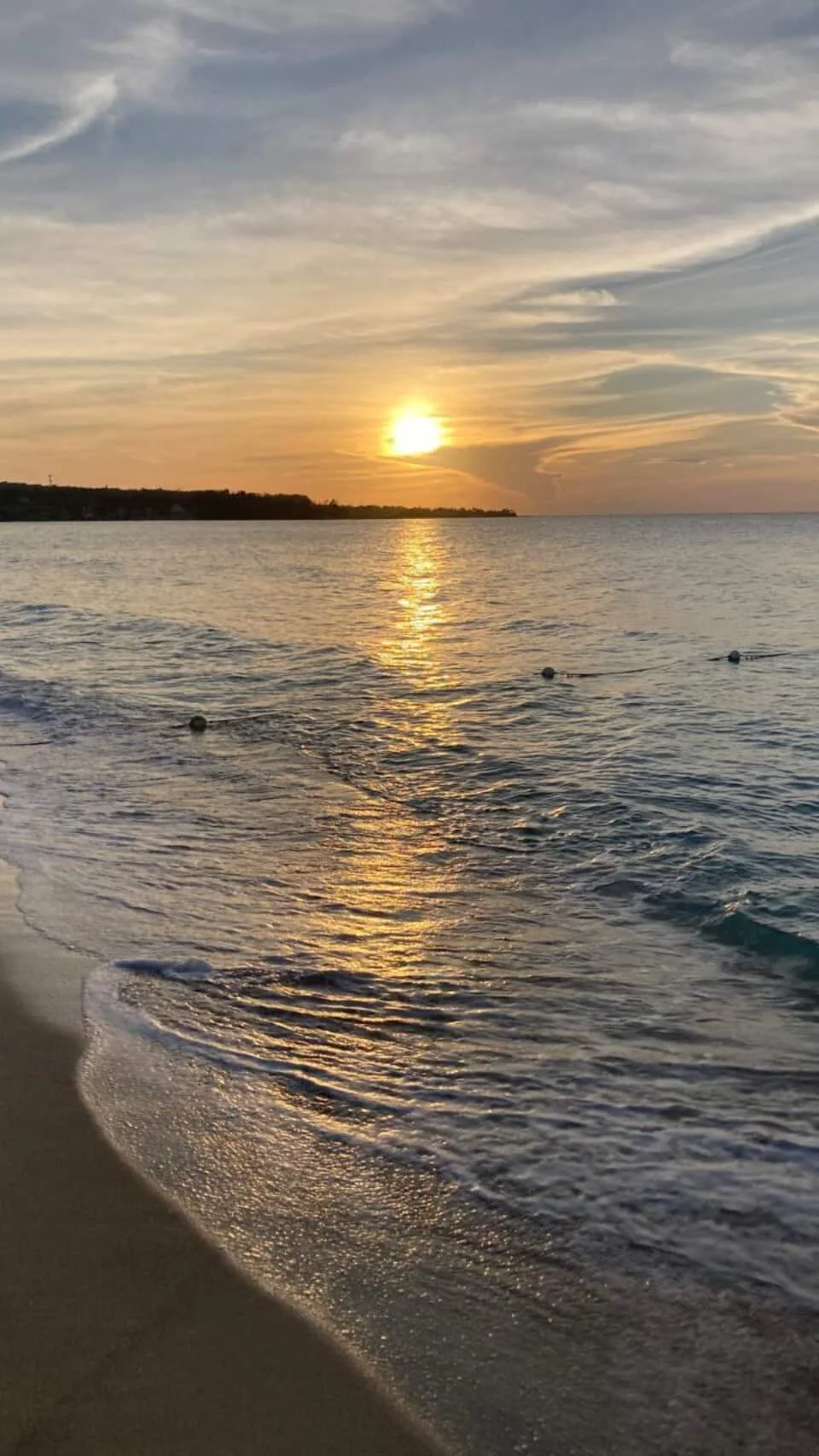 Sunset over calm ocean waters with a sandy beach in the foreground, and buoys floating on the surface.