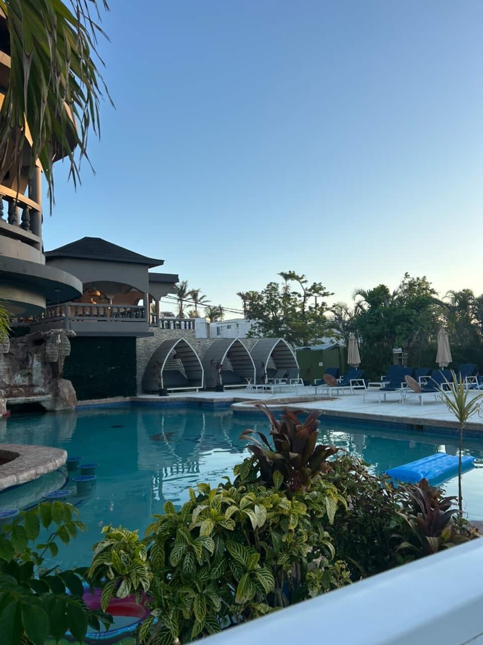 A swimming pool at a resort with lounge chairs and umbrellas around it, surrounded by lush green trees and tropical plants under a clear blue sky.