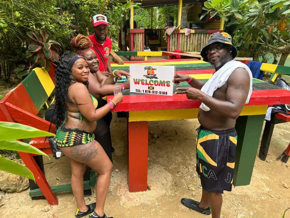 Four people, three women and one man, are gathered around a colorful outdoor table, with a welcome sign on it. The women are dressed in swimsuits and the man is shirtless with a towel over his shoulder. They are in a lush, green tropical setting, smi