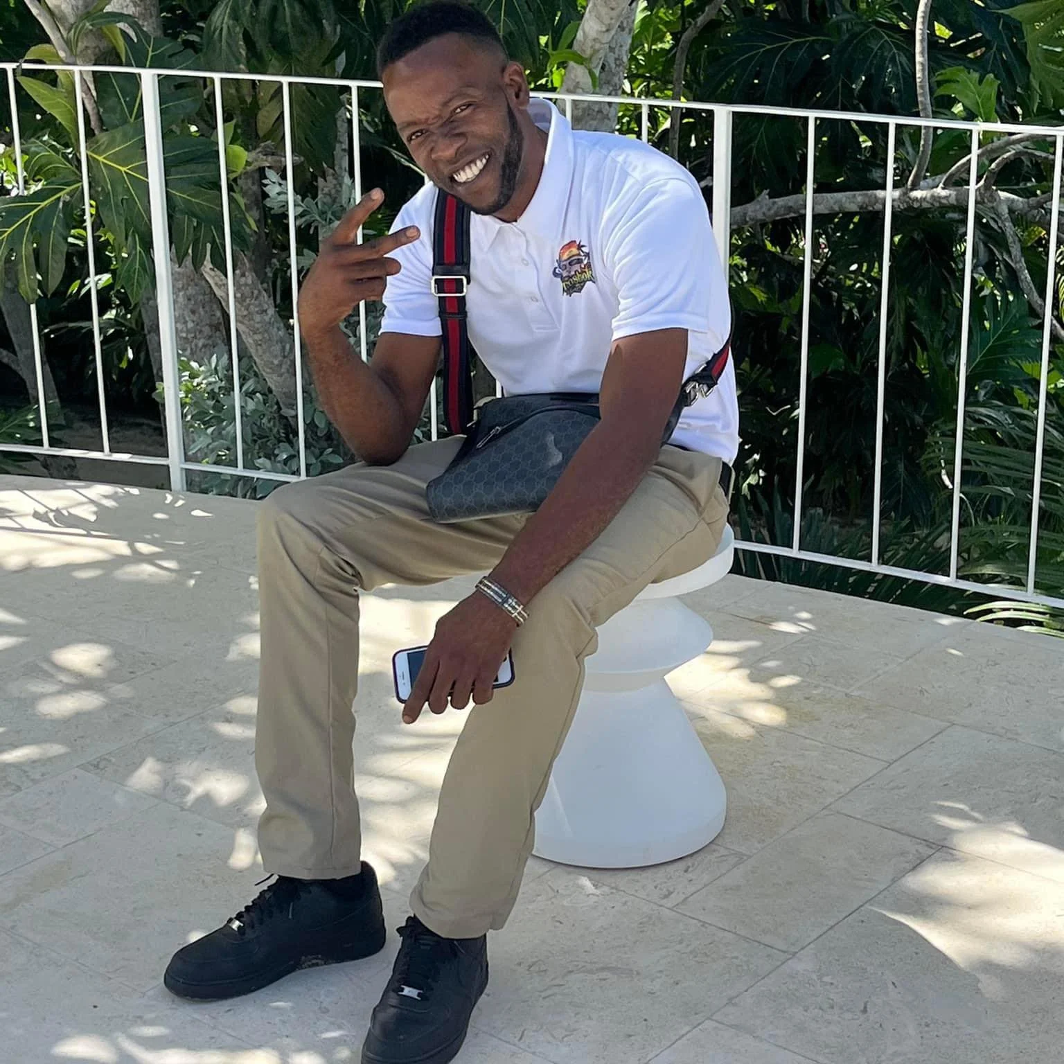 A man sitting on a white toilet outdoors, smiling and making a peace sign with his right hand. He is wearing a white polo shirt, beige pants, and black shoes, with a black crossbody bag. He is holding a smartphone in his left hand, and there is greenery and a white metal fence in the background.