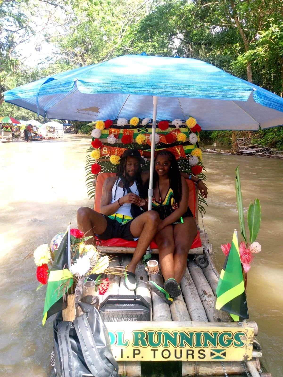 Two people sitting on a bamboo raft decorated with flags and flowers, under a blue umbrella, on a river surrounded by trees, with a sign reading 'Cool Runnings U.P. Tours'.