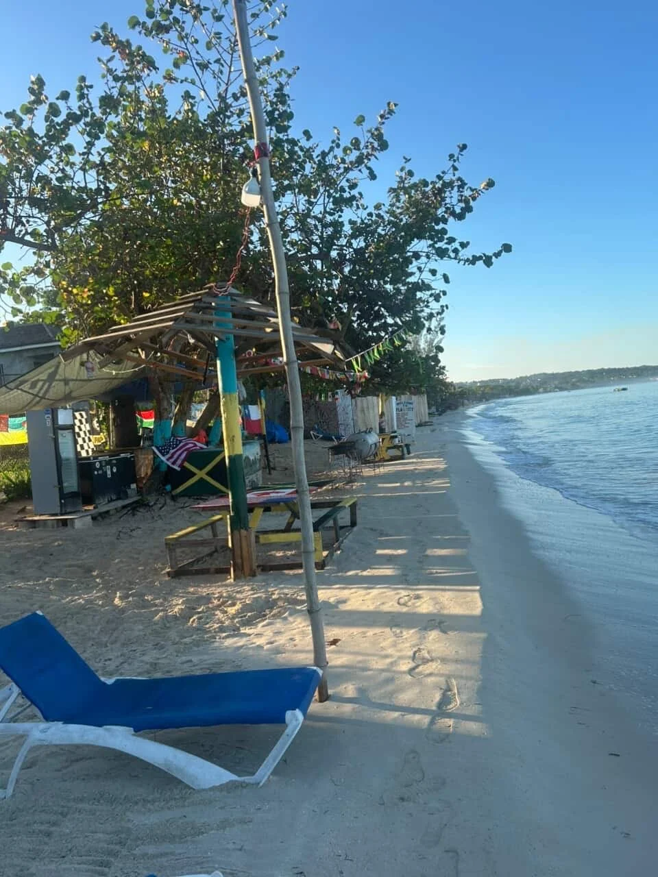 Empty beach with a blue lounge chair, umbrellas, and small structures along the shoreline, with trees and a clear sky in the background.