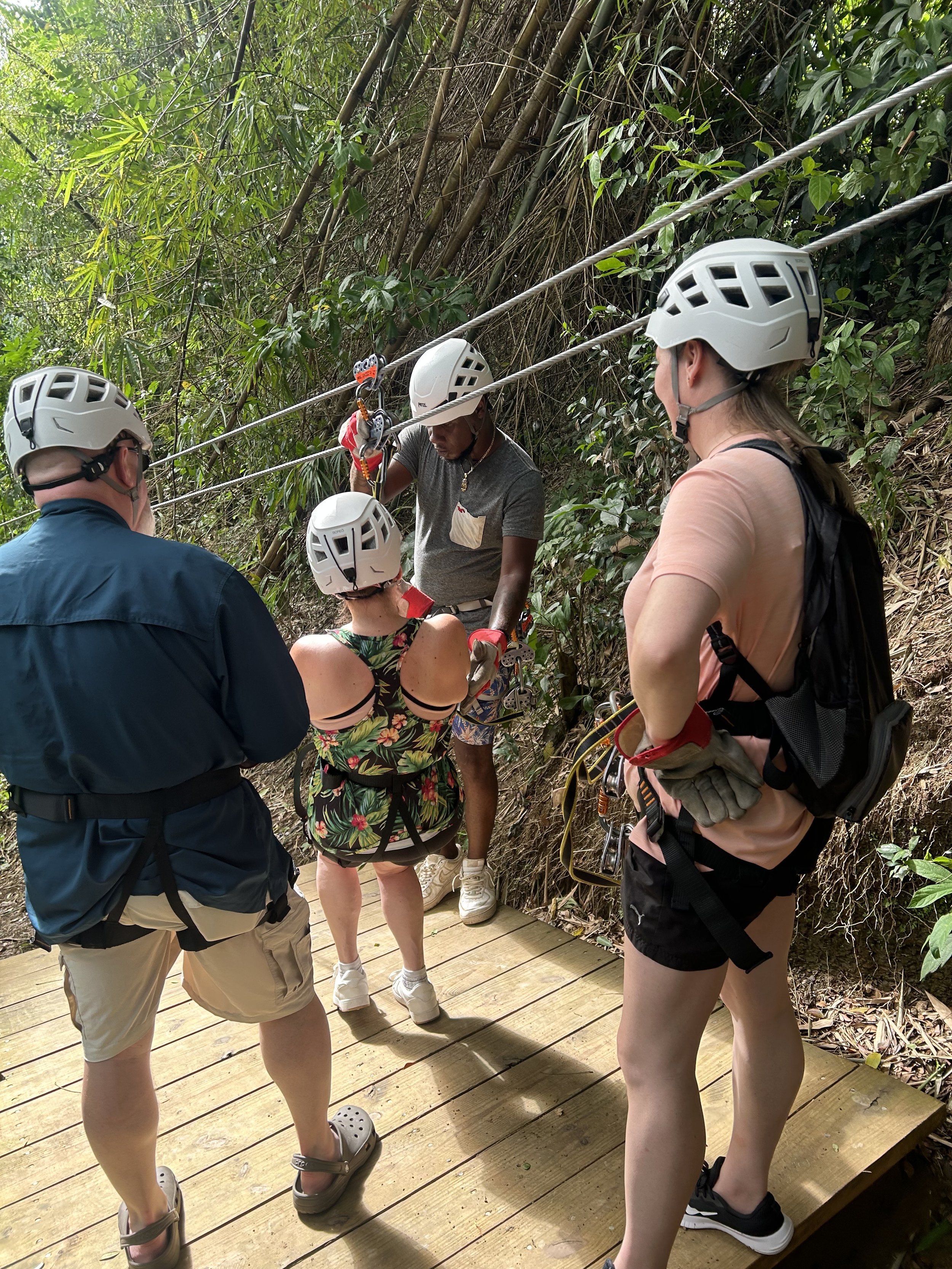 Group of people preparing for zip-lining on a forested platform, wearing helmets and harnesses.