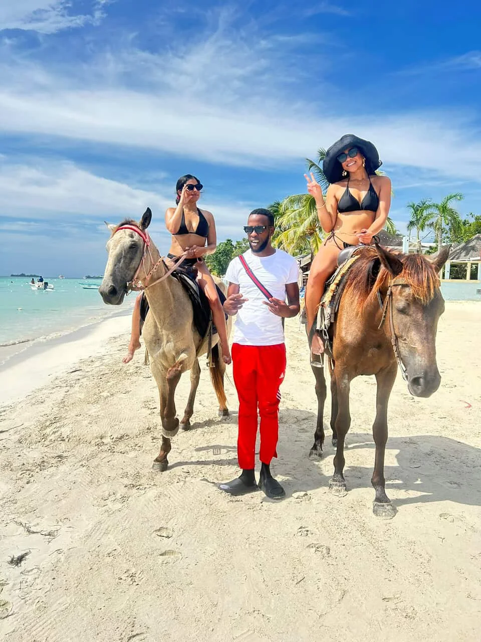 Three people at the beach, two women riding horses and a man standing in between them, with a backdrop of blue sky, ocean, and palm trees.