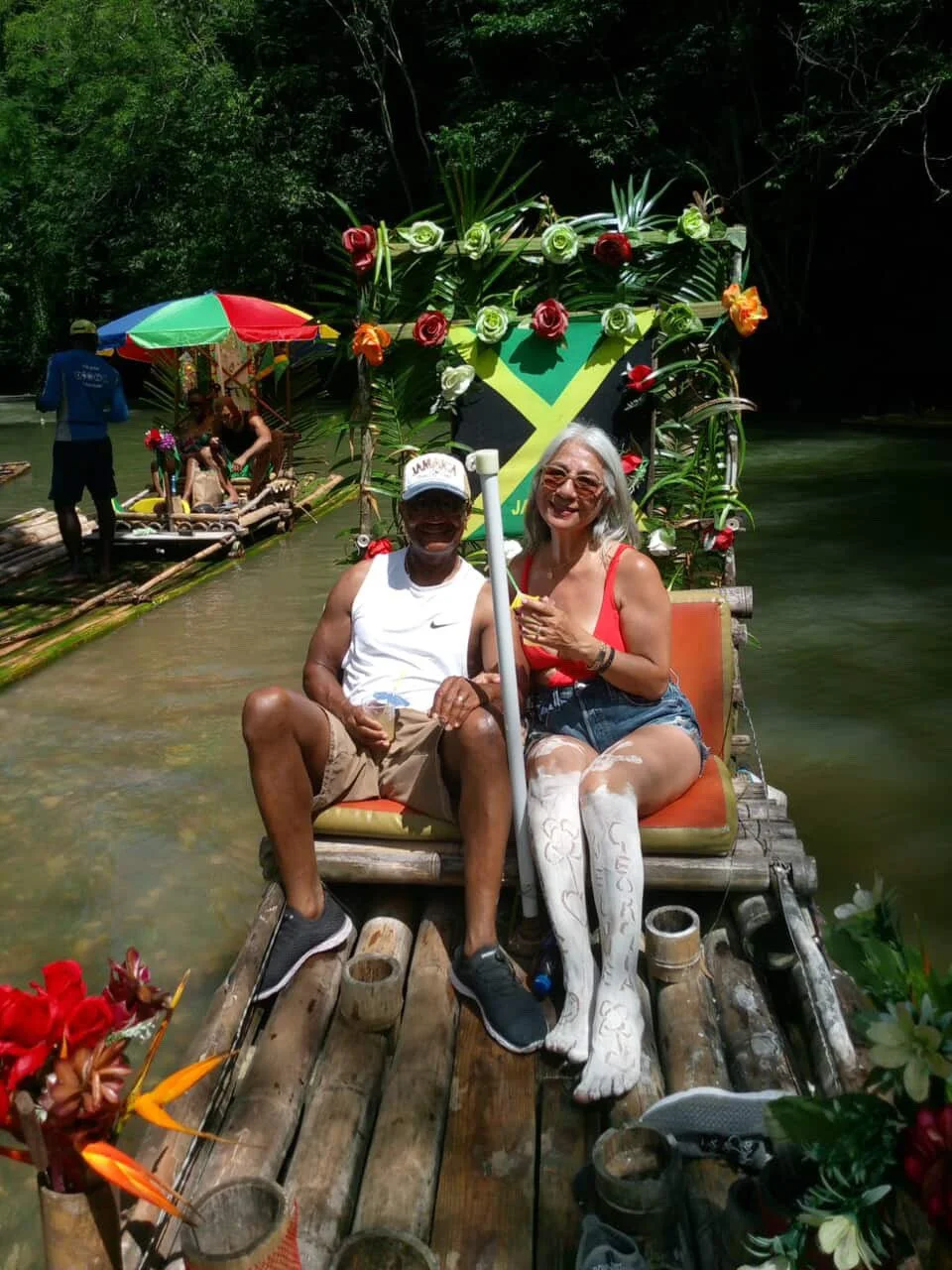 Two people sitting on a bamboo raft decorated with tropical flowers, with a flag of Jamaica in the background. They are smiling and enjoying a water-based activity in a forested area.