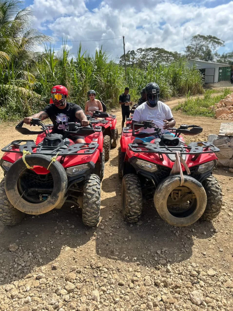 Four people wearing helmets on red all-terrain vehicles (ATVs) on a dirt path in a rural area with green plants and a blue sky.