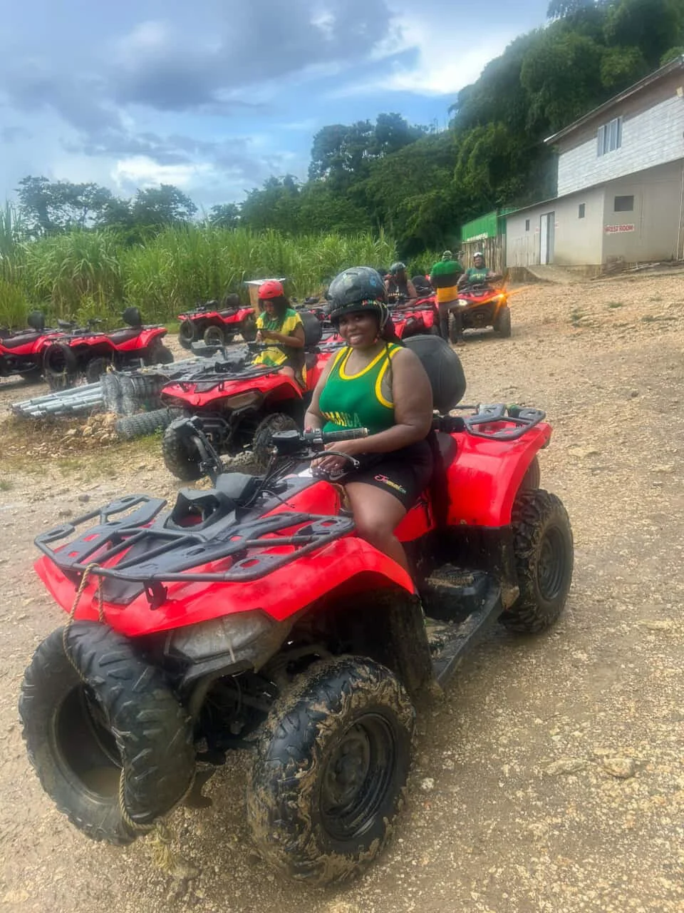 Group of women riding all-terrain vehicles (ATVs) on a dirt path, with some ATV riders standing in the background, greenery, and a building nearby.