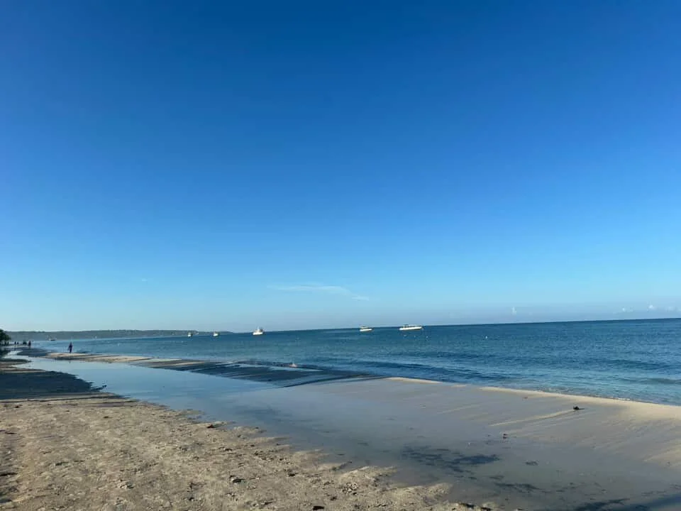 A calm beach with sandy shoreline, clear blue water, and a bright blue sky with a few clouds.