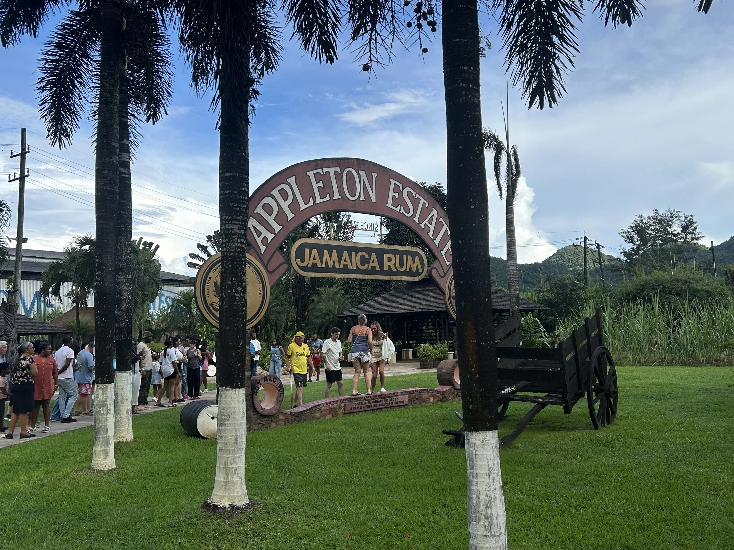 Entrance sign for Appleton Estate Jamaica Rum with a group of people waiting in line, surrounded by palm trees and lush greenery, under a cloudy sky.