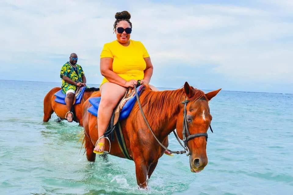 Two people horseback riding in the water at the beach, one woman in a yellow shirt and dark sunglasses, the other man in a colorful shirt and sunglasses, both on chestnut-colored horses.
