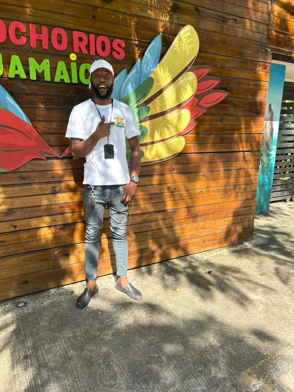 A man standing outdoors in front of a wooden wall with colorful text that says 'Pochorios Jamaica' and a graphic of a wing with yellow, green, red, and blue feathers. The man is wearing a white cap, white t-shirt, gray jeans, and gray shoes, and is p