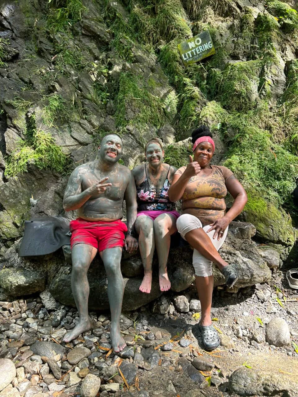 Three people sitting on rocks near a mossy rock wall, covered in mud and smiling. One person is giving a thumbs-up, another making a peace sign, and the third raising a fist. A sign above reads 'No Littering'.
