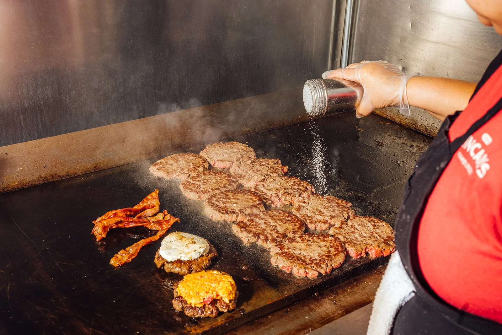 A cook seasoning hamburger patties on a flat grill with salt, with bacon, two cooked eggs, and additional burger patties around.