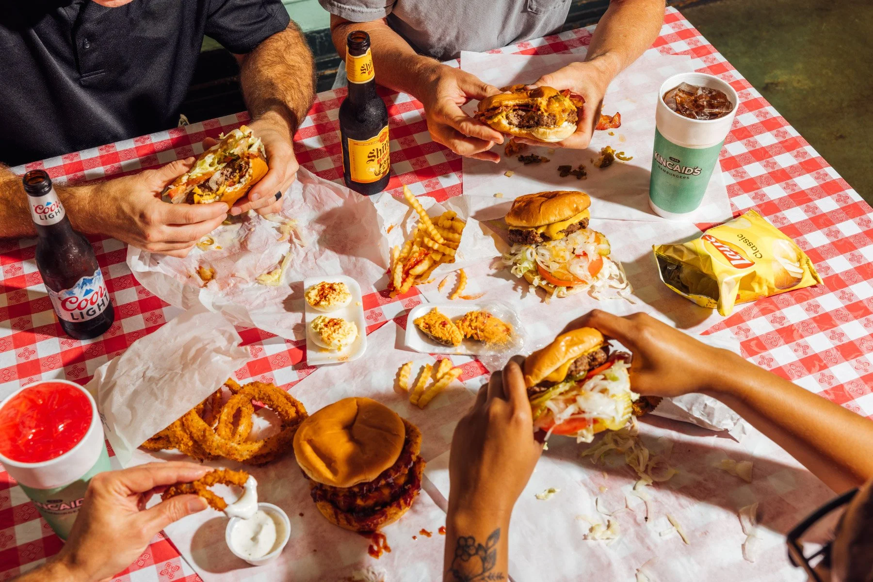 People enjoying a casual meal with cheeseburgers, fries, onion rings, and drinks on a red and white checkered tablecloth.