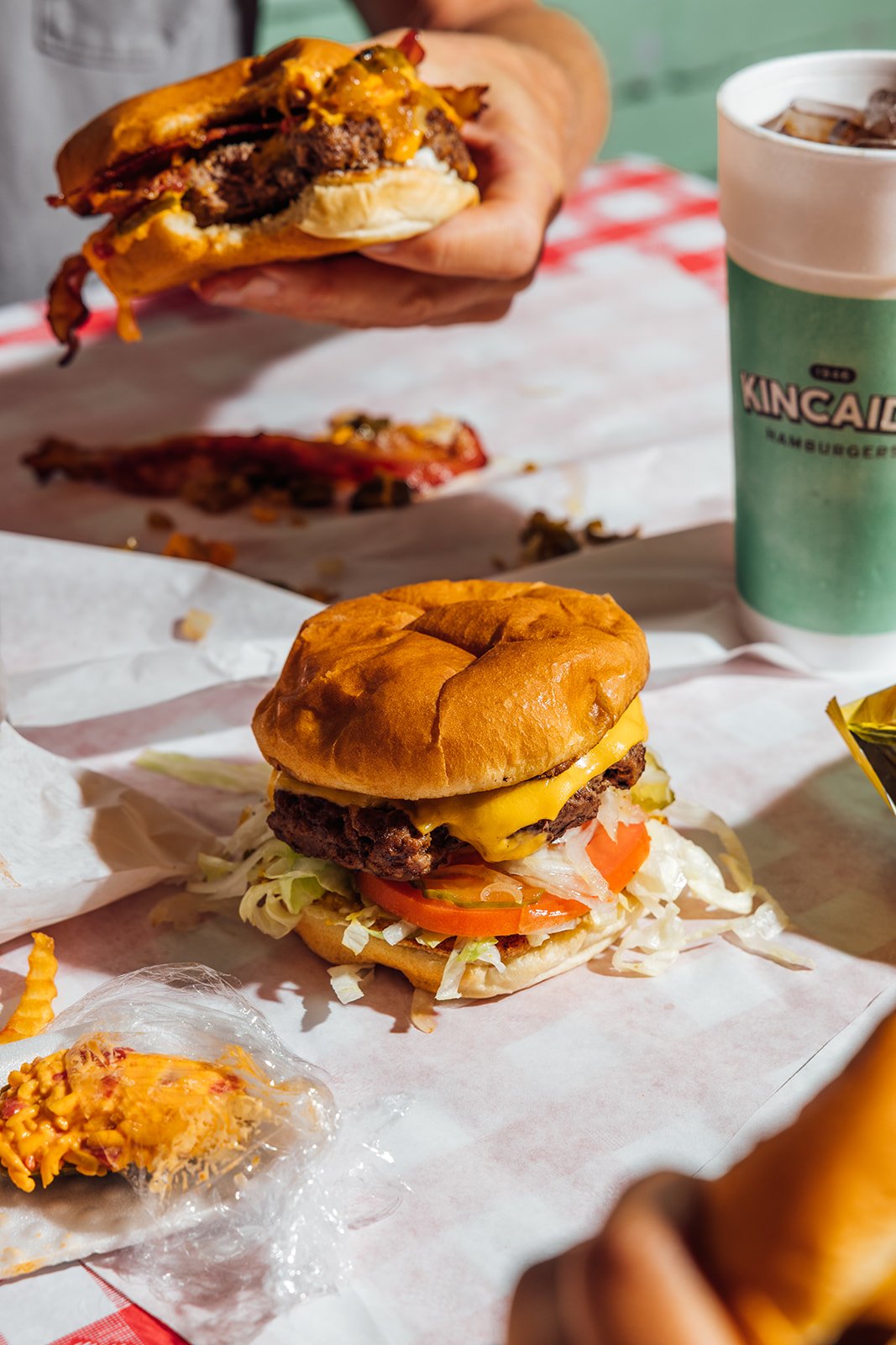 A cheeseburger with lettuce, tomato, pickles, and a beef patty, topped with melted cheddar cheese on a bun, placed on white paper. In the background, another burger and a large soda cup are visible, with a red-checkered tablecloth.