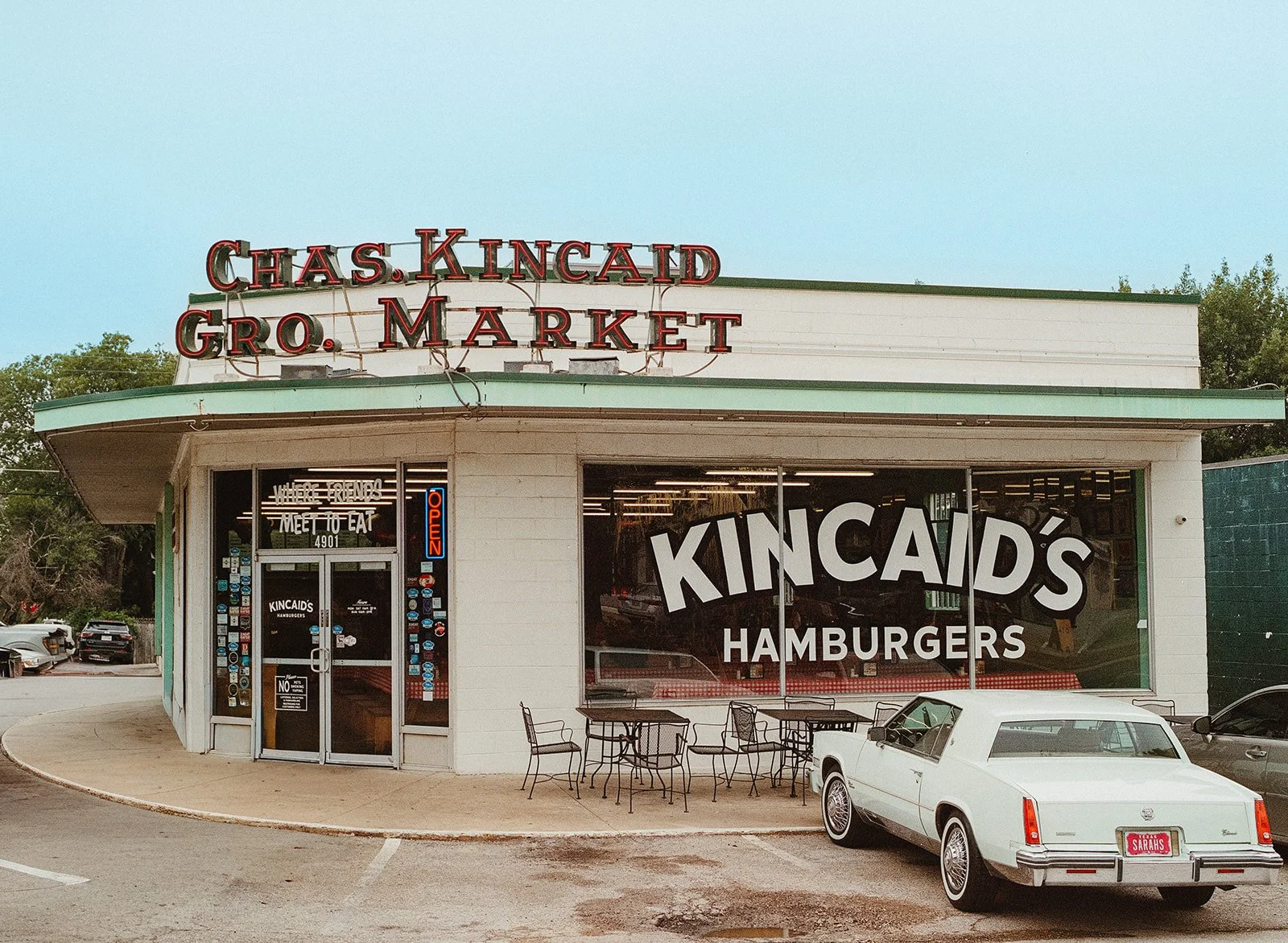 Vintage Kincaid's Hamburgers restaurant with a white exterior, large front window, outdoor seating, neon 'Open' sign, and classic car parked outside.